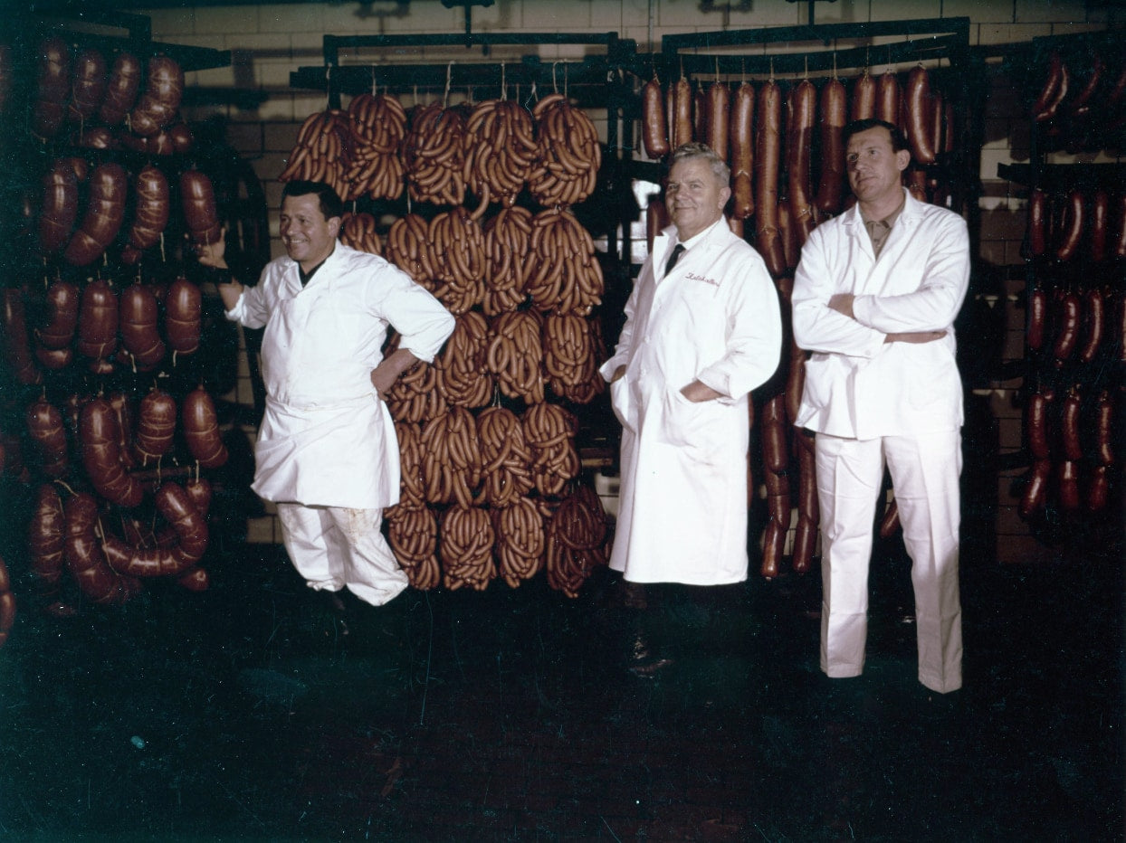 Vintage photo of Schaller Weber and two employees in production facility with hanging strings of sausages and salami.